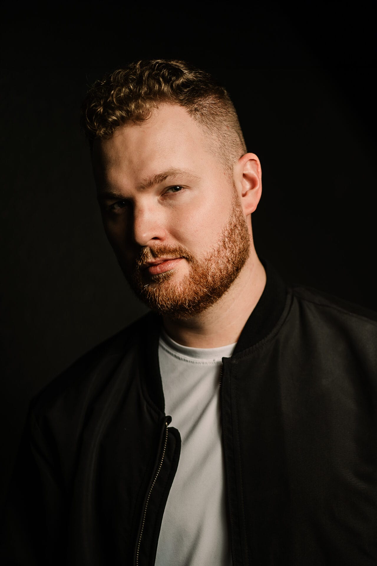 Phoenix Wedding Photographer 8 Portrait of a phoenix wedding photographer with curly hair and a beard, wearing a black bomber jacket over a white shirt, set against a dark background. The image highlights his facial features and confident expression.