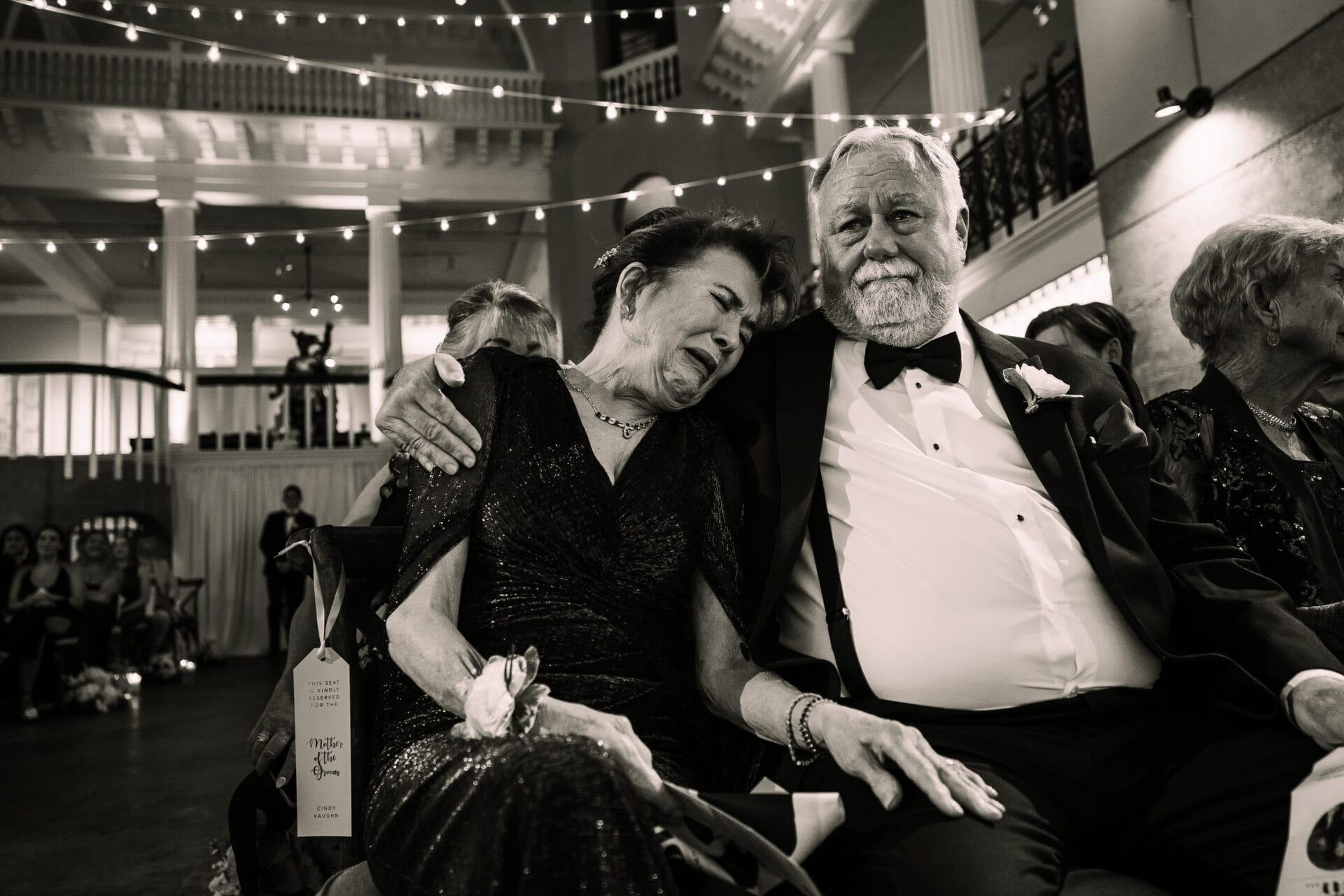 Phoenix Wedding Photographer 7 Emotional moment at a wedding with an elderly couple seated together, the woman is visibly emotional, and the man gently comforts her. The setting features warm string lights and elegant decor, capturing the intimate atmosphere of the celebration. A decorative tag is visible on the chair, adding a personal touch.
