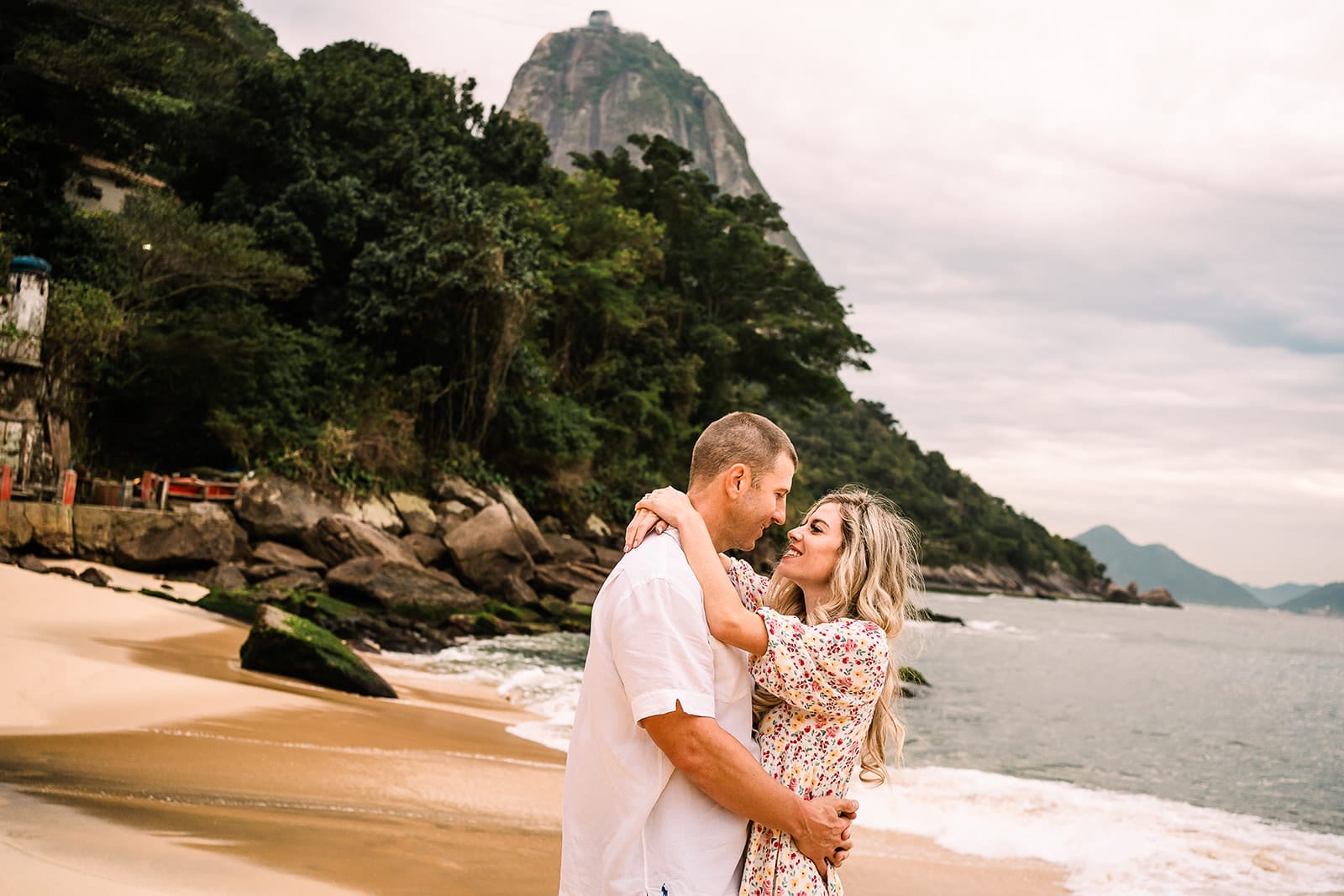 Best Things To Do in Rio de Janeiro for Couples in 2025 2 Couple embracing on a beach with rocky coastline and mountains in the background, overcast sky, showcasing romantic atmosphere and scenic views.