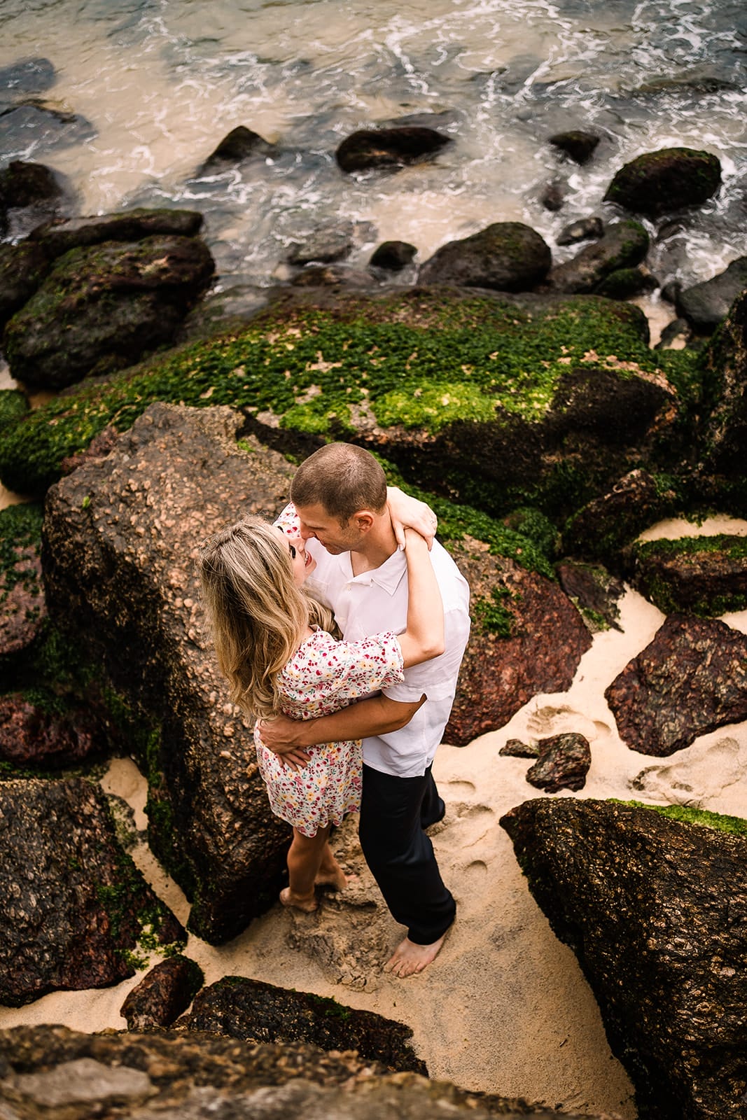 Couple embracing on a beach surrounded by rocks and sand, with gentle ocean waves in the background. The woman wears a floral dress, while the man is in a white shirt, creating a romantic outdoor setting.