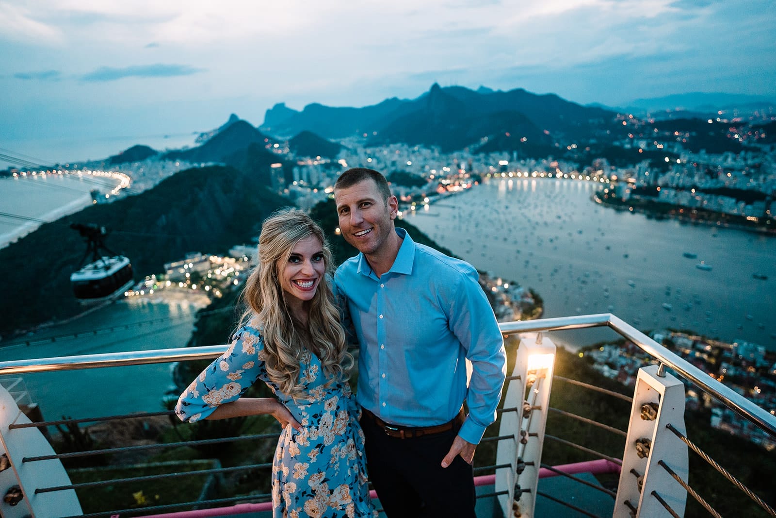 Best Things To Do in Rio de Janeiro for Couples in 2025 10 Couple posing happily at a scenic viewpoint in Rio de Janeiro, with a cityscape and coastline in the background; the sun sets, illuminating the area with city lights and a gondola in the distance.