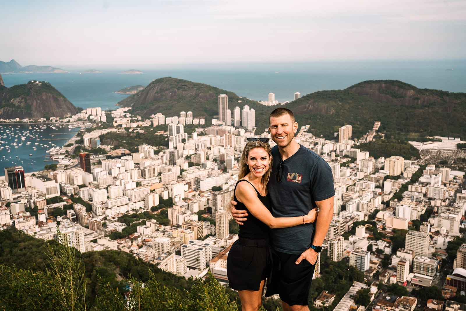 Best Things To Do in Rio de Janeiro for Couples in 2025 4 Couple posing with a cityscape view of Rio de Janeiro, Brazil, featuring buildings, mountains, and a marina in the background. The image captures a scenic landscape with the ocean visible in the distance.