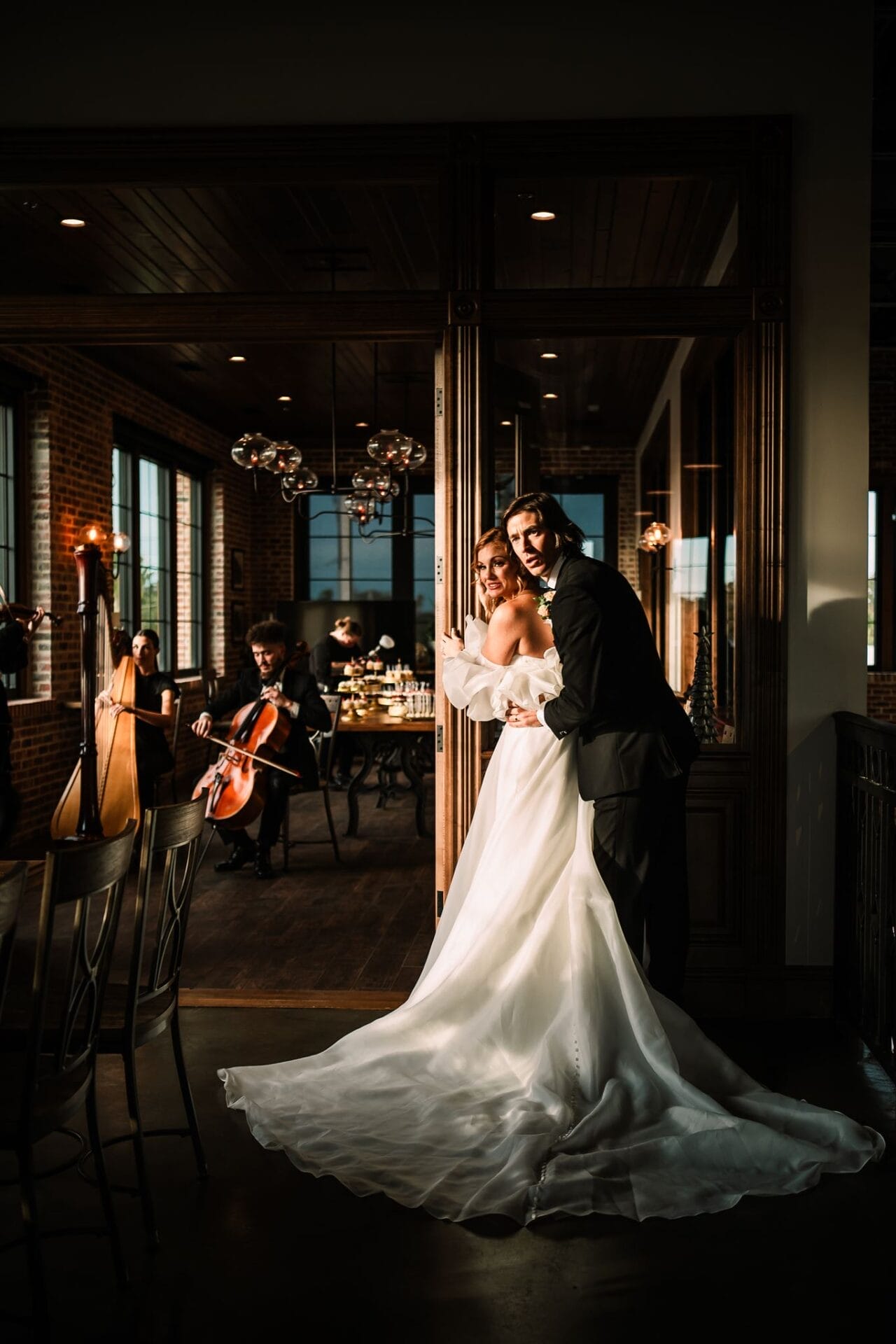 Phoenix Wedding Photographer 6 Bride and groom embrace at the entrance of a stylish venue, featuring dim lighting, rustic brick walls, and a string quartet playing in the background. The bride wears a flowing white gown while the groom is in a black suit.
