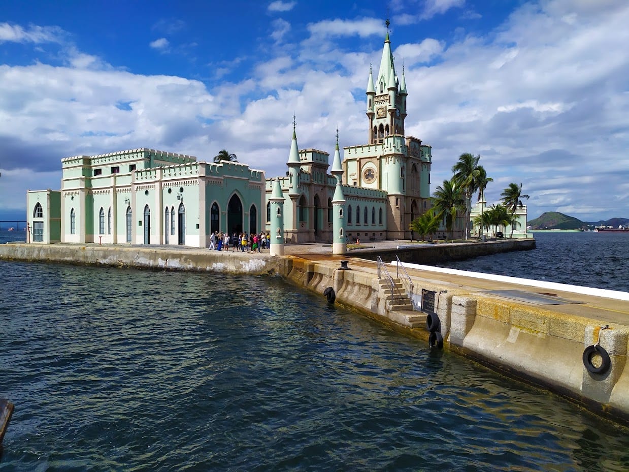 Historic castle by the waterfront with a green facade, featuring a clock tower and palm trees. Visitors gather near the dock, overlooking the ocean under a partly cloudy sky.