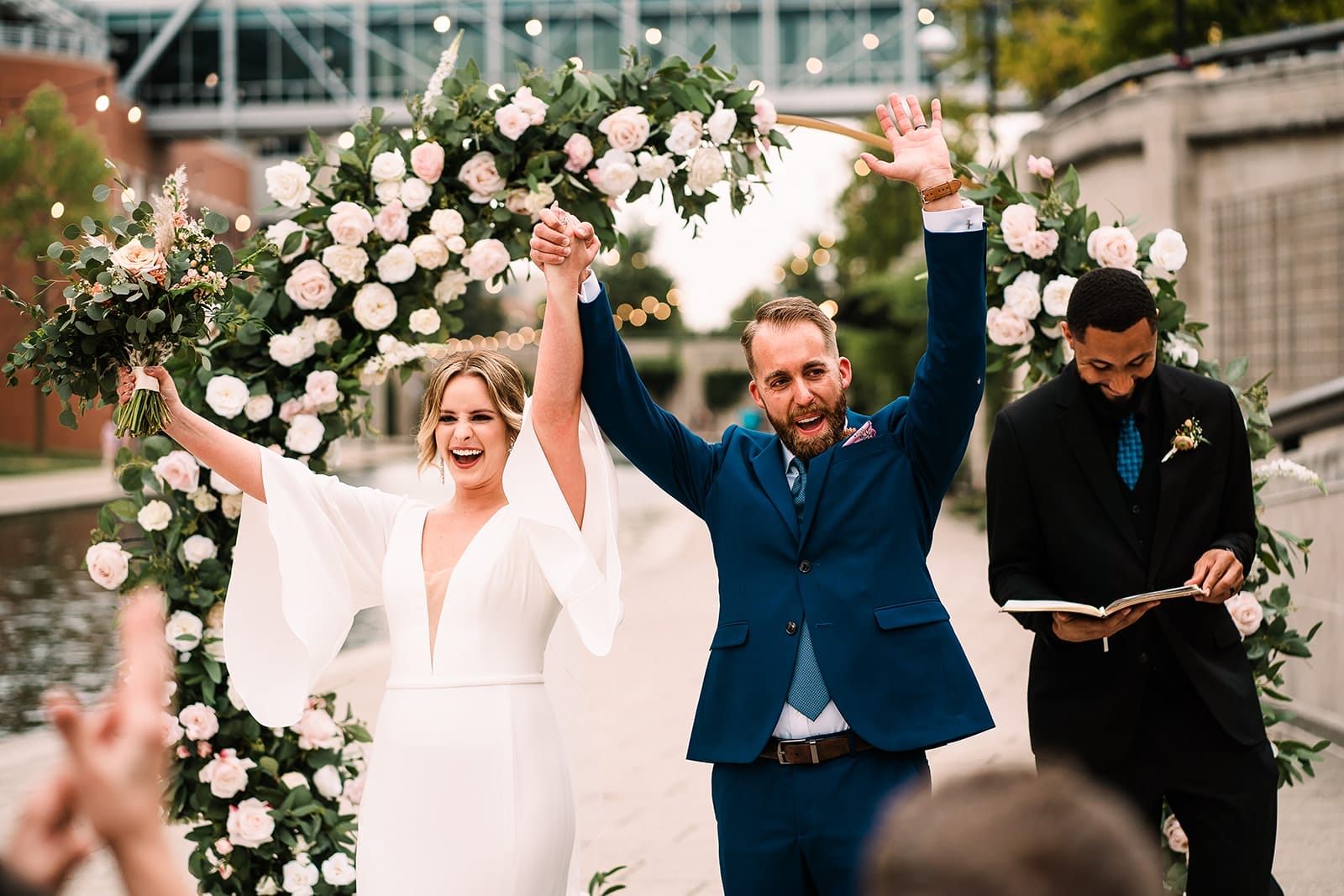 Bride and groom celebrating their wedding ceremony, smiling and raising their hands in joy. The bride wears a stylish white dress and holds a bouquet, while the groom is in a navy suit. An officiant stands nearby, reading from a book, framed by a floral archway adorned with white and pink roses.
