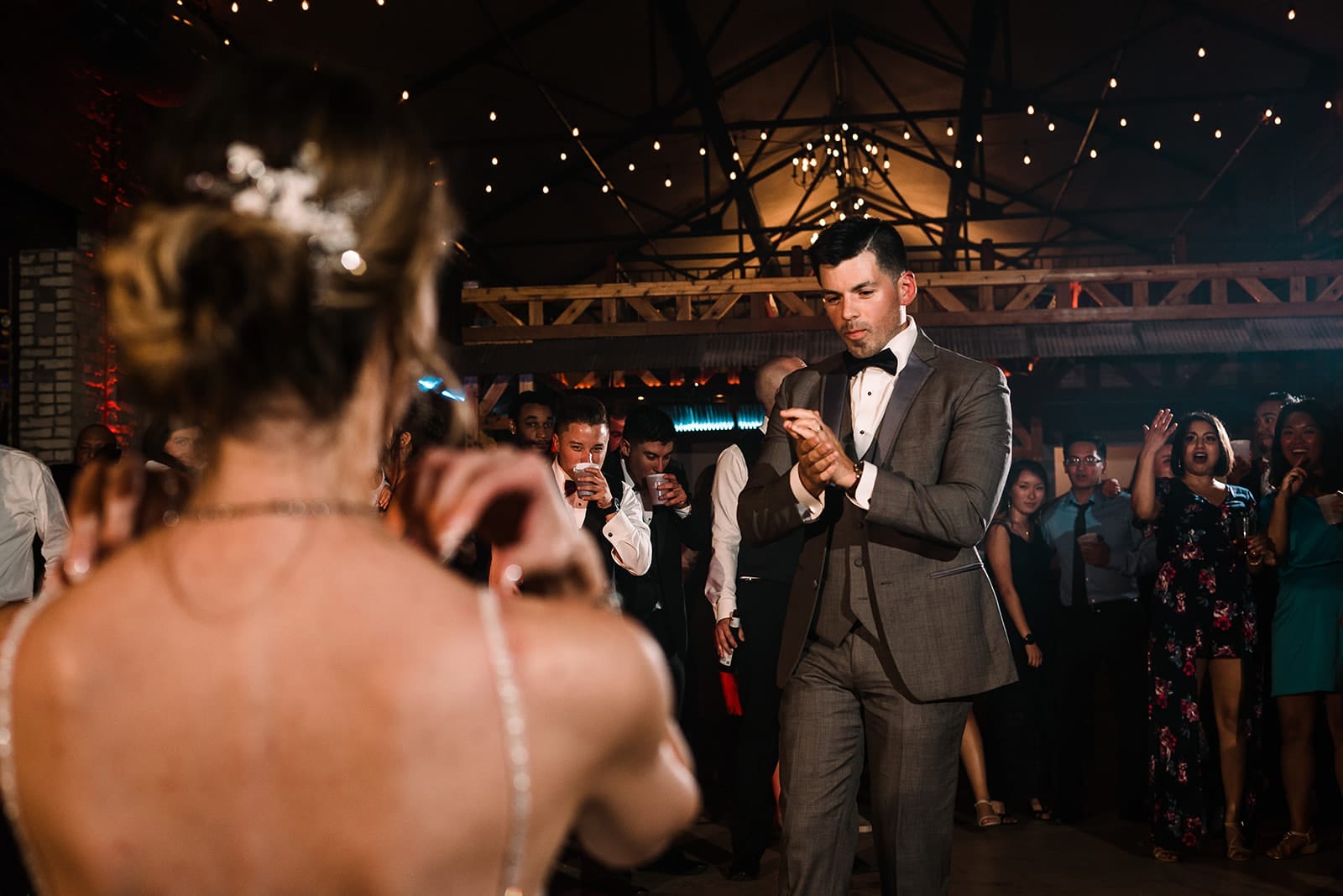 A stylish man in a gray suit and bow tie dances at a lively event, surrounded by guests enjoying the celebration. The scene features warm lighting and a festive atmosphere, with attendees clapping and engaging in the moment.