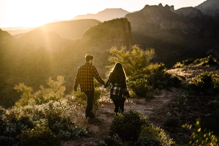Couple walking hand in hand along a rocky trail during sunset, surrounded by greenery and mountain scenery.