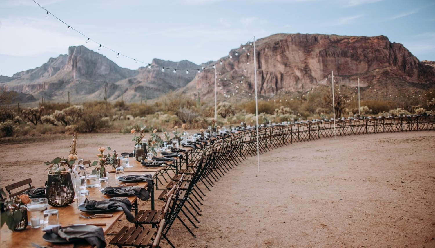 Outdoor dining setup with a long wooden table and metal chairs surrounded by desert scenery and mountains. The table is elegantly decorated with flowers and glassware, ideal for events or gatherings. String lights are installed overhead, enhancing the rustic ambiance.
