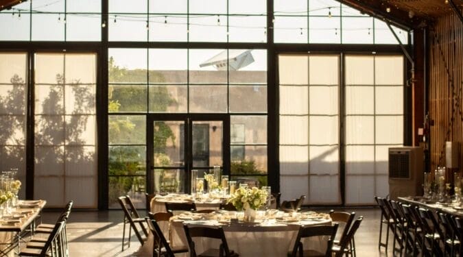 Best Small Wedding Venues in Phoenix, AZ (Intimate & Micro Weddings) 7 Elegant event space featuring round tables set for a formal gathering, decorated with floral centerpieces. Large glass windows allow natural light to illuminate the interior, providing a scenic view of greenery outside. The setting is ideal for weddings or corporate events.