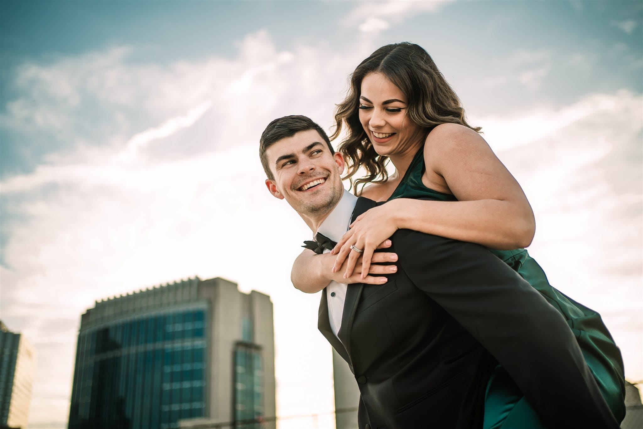 Couple enjoying a playful moment outdoors, with the man giving the woman a piggyback ride. Both are dressed elegantly, with a city skyline and a clear sky in the background.