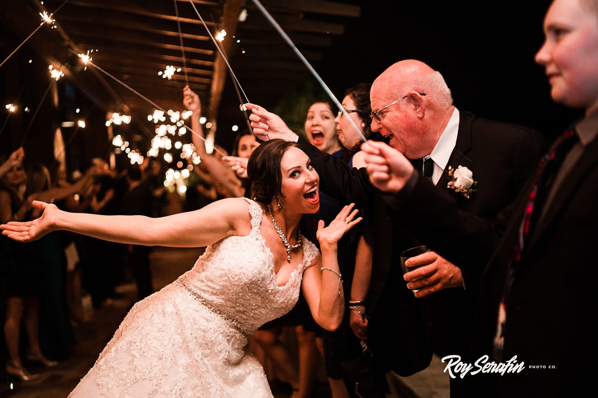 Bride celebrating with guests at a wedding reception, surrounded by sparklers and joyful expressions. The bride is in a detailed white gown, posing energetically with her arm extended. Friends and family are cheering in the background, creating a festive atmosphere.