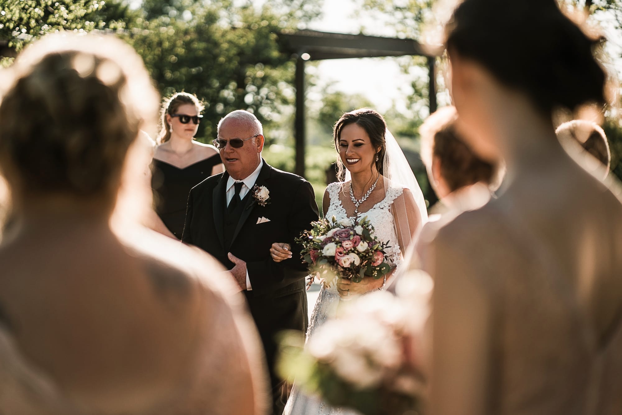 Bride walking down the aisle with her father at an outdoor wedding ceremony, surrounded by bridesmaids and guests. The bride holds a bouquet of flowers and wears a lace gown and veil, while her father is in a formal suit. The scene is set in a lush garden with soft sunlight filtering through the trees.