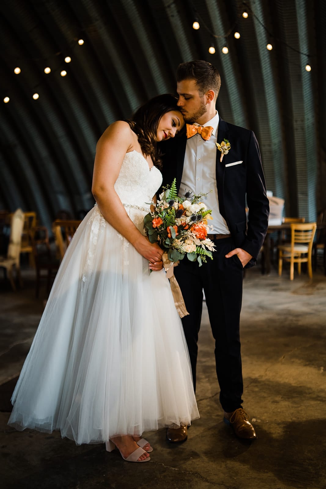 Bride and groom posing together in a rustic venue, the bride wearing a lace wedding gown and holding a floral bouquet, while the groom is dressed in a black suit with an orange bow tie. Soft lighting enhances the romantic atmosphere.