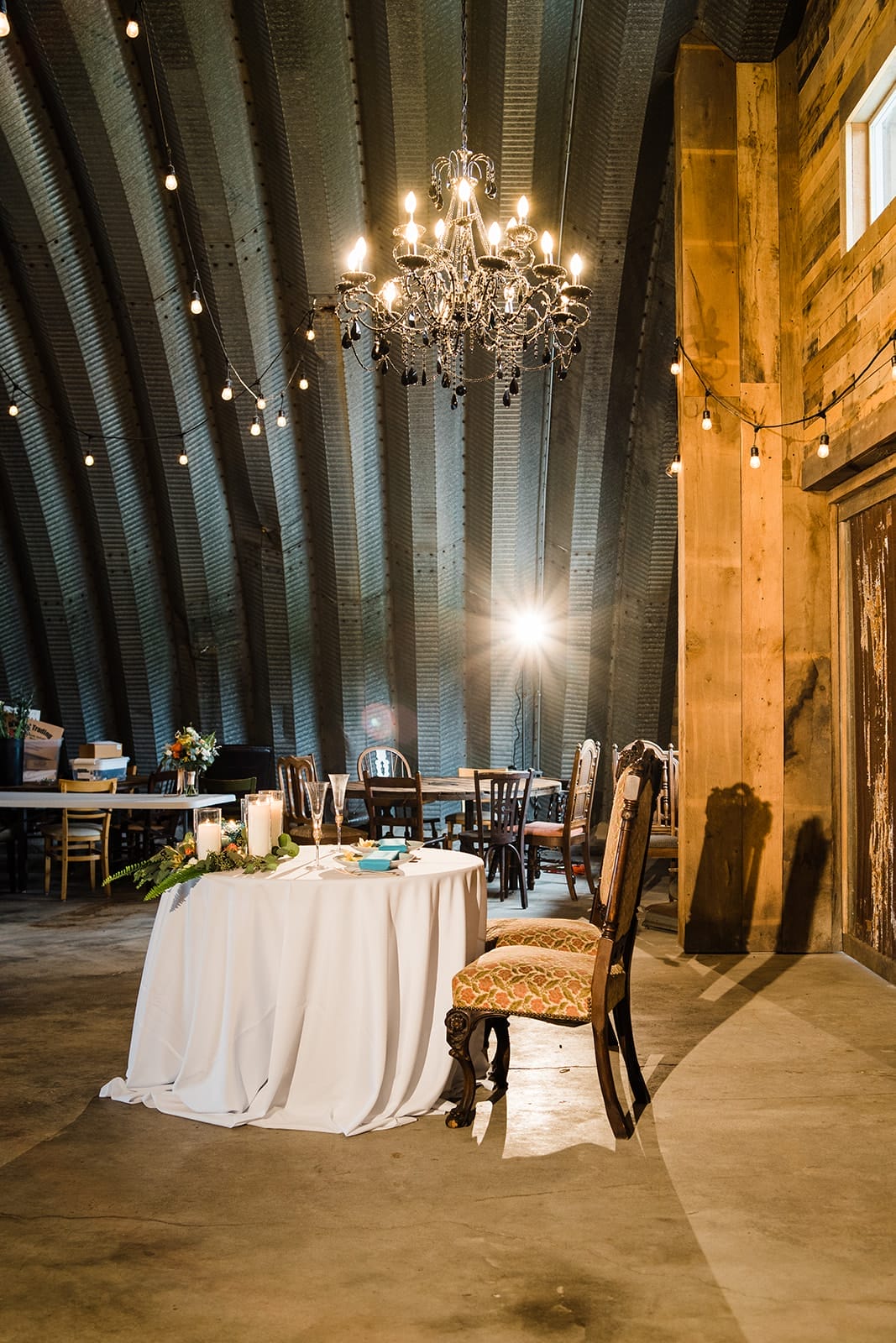 Elegant event setting featuring a white tablecloth table adorned with candles and floral arrangements, a vintage chair, and a chandelier under a curved metal ceiling. The background showcases additional rustic wooden furniture, creating a warm atmosphere ideal for gatherings or celebrations.