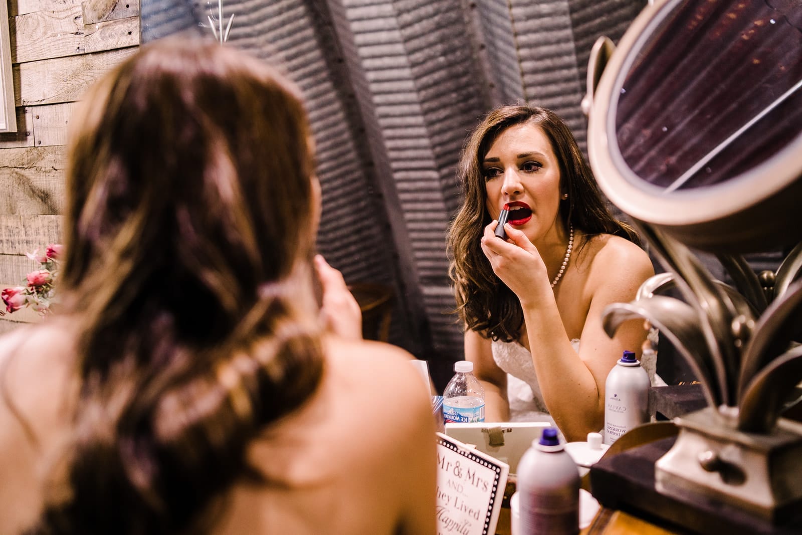 Bride applying red lipstick in front of a mirror, preparing for her wedding day, with floral decor and grooming products visible in the background.