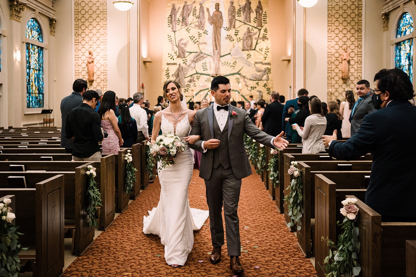Bride and groom walking down the aisle after their wedding ceremony in a church, surrounded by guests celebrating. The bride holds a bouquet of flowers and wears a lace wedding dress, while the groom is in a formal suit with a bow tie. The interior features stained glass windows and floral decorations along the pews.