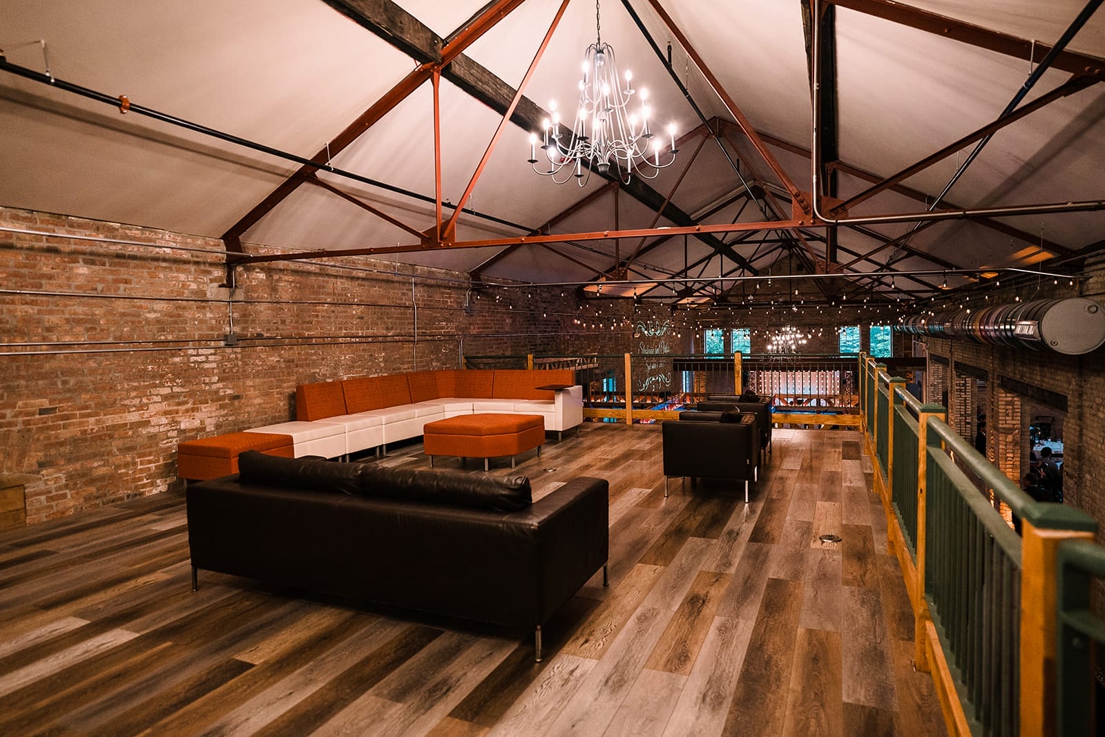 Spacious loft interior featuring modern furniture, including a white and orange sectional sofa and black leather couches, illuminated by a stylish chandelier. The space has exposed brick walls, wooden flooring, and large windows with string lights for a cozy ambiance, making it ideal for gatherings or events.