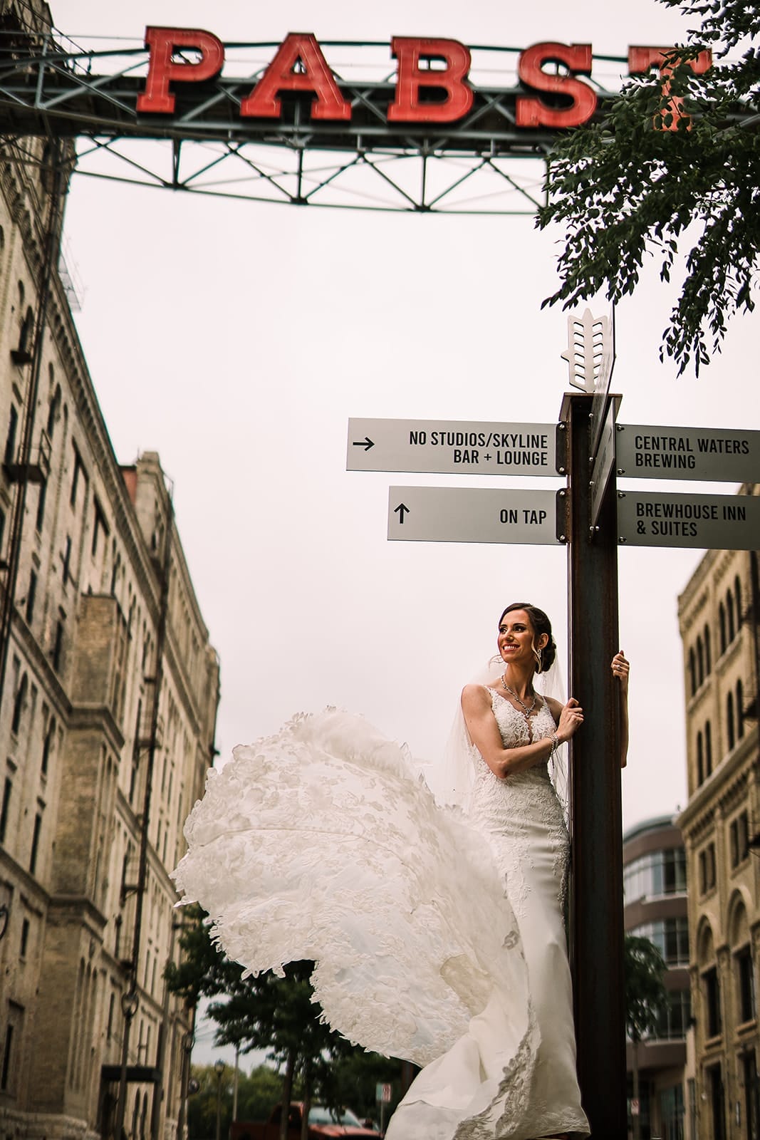 Bride in a flowing wedding dress poses at a directional sign under the Pabst sign in an urban setting. The scene captures a blend of modern and historic architecture, with notable landmarks such as Central Waters Brewing and Brewhouse Inn & Suites visible on the sign.
