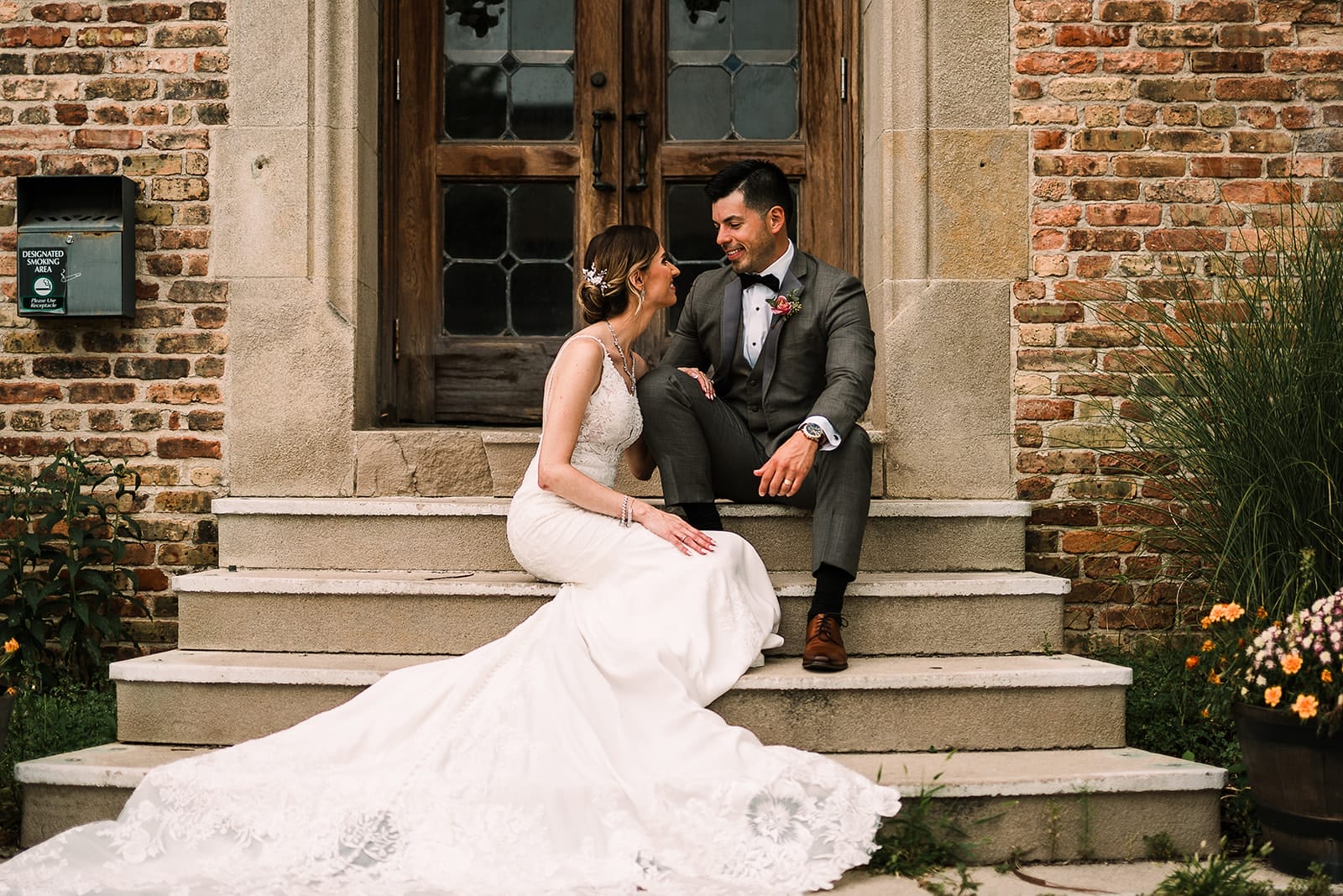 Bridal couple sitting together on stone steps in front of a brick building, smiling and looking at each other. The bride wears a lace wedding gown, while the groom is in a gray suit with a bow tie. Floral decorations and greenery surround the location, contributing to a romantic atmosphere.