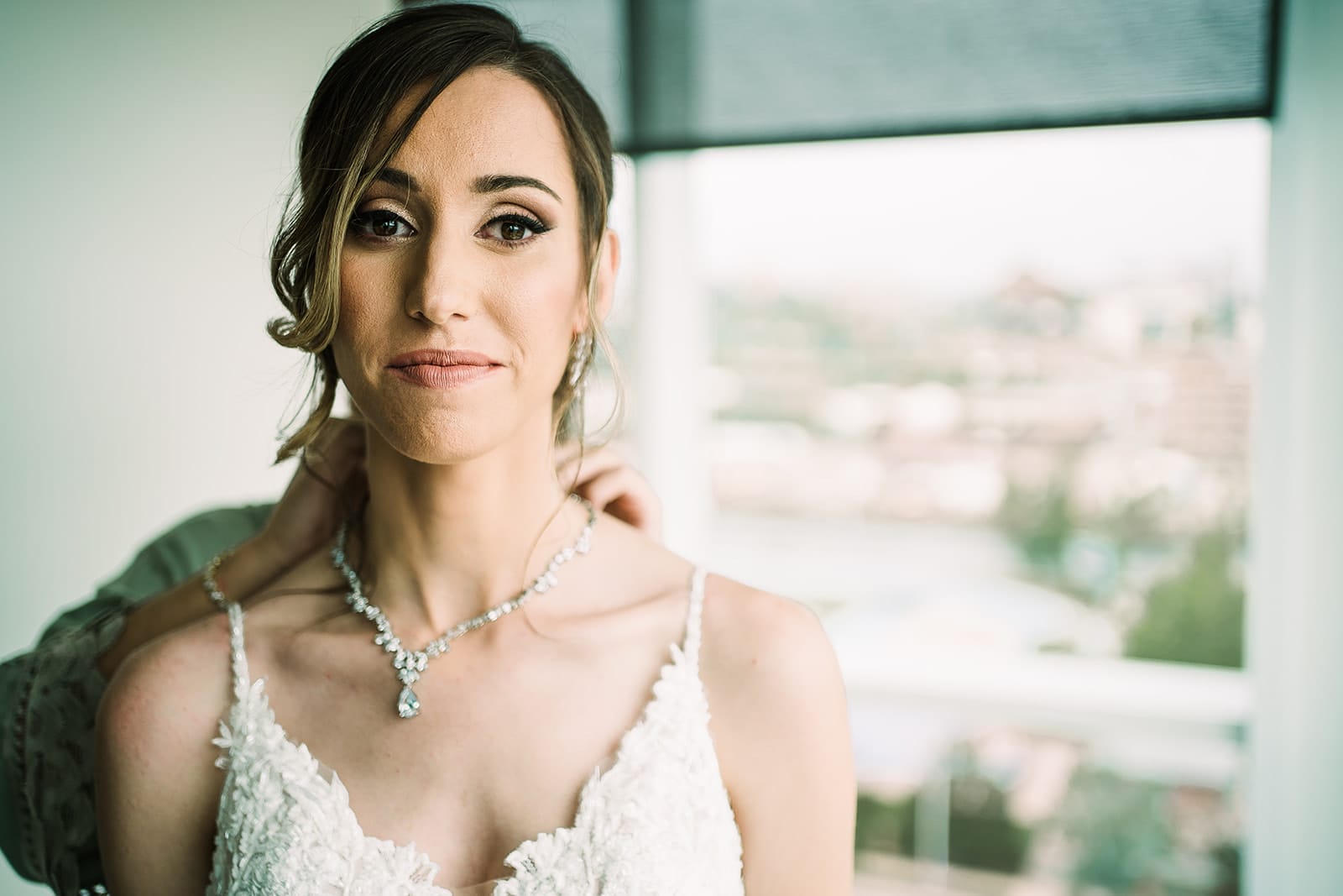 Bride getting ready in a bridal gown, wearing a sparkling necklace, with a soft makeup look, as another person adjusts her jewelry. Background features a blurred view of a city.