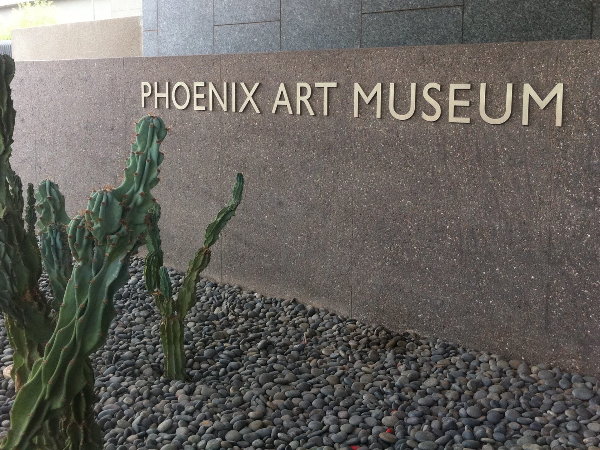 Signage of the Phoenix Art Museum displayed on a stone wall, with cacti and smooth pebbles in the foreground.