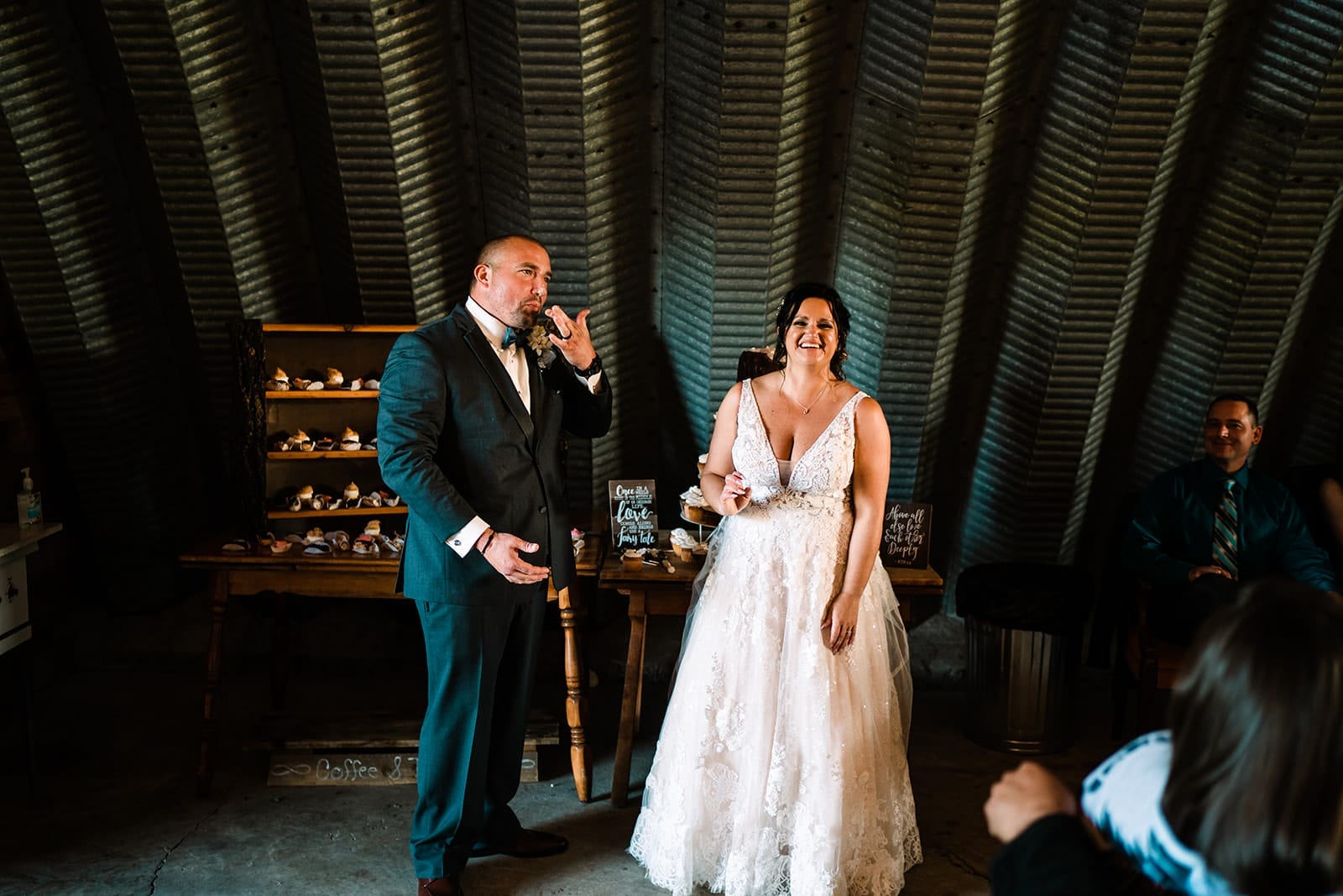 Bride and groom laughing together during their wedding reception, surrounded by guests in a rustic venue. The groom is making a playful gesture while the bride, wearing a lace wedding dress, smiles joyfully. A dessert table is visible in the background, enhancing the celebratory atmosphere.