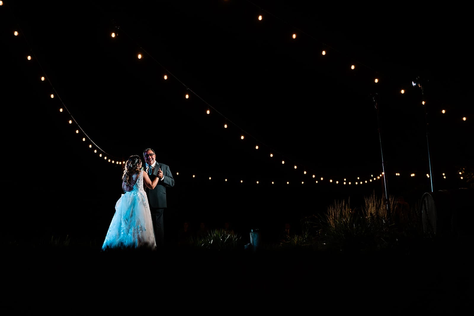 Couple dancing together under string lights at a nighttime wedding reception, surrounded by a dark sky and greenery.