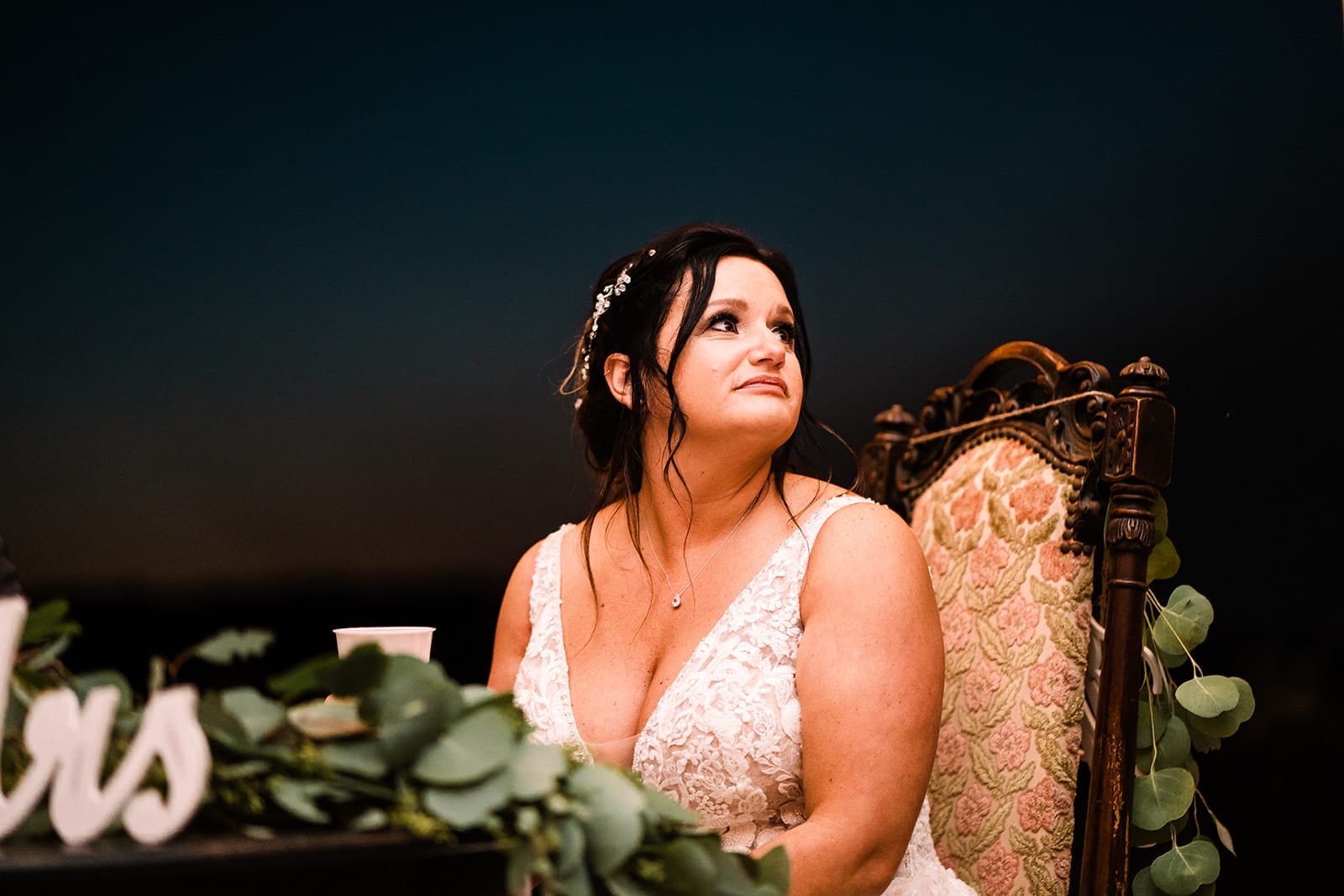 Bride sitting at a decorated reception table, wearing an elegant lace wedding dress and looking thoughtfully into the distance, with greenery in the foreground and a dark evening background.