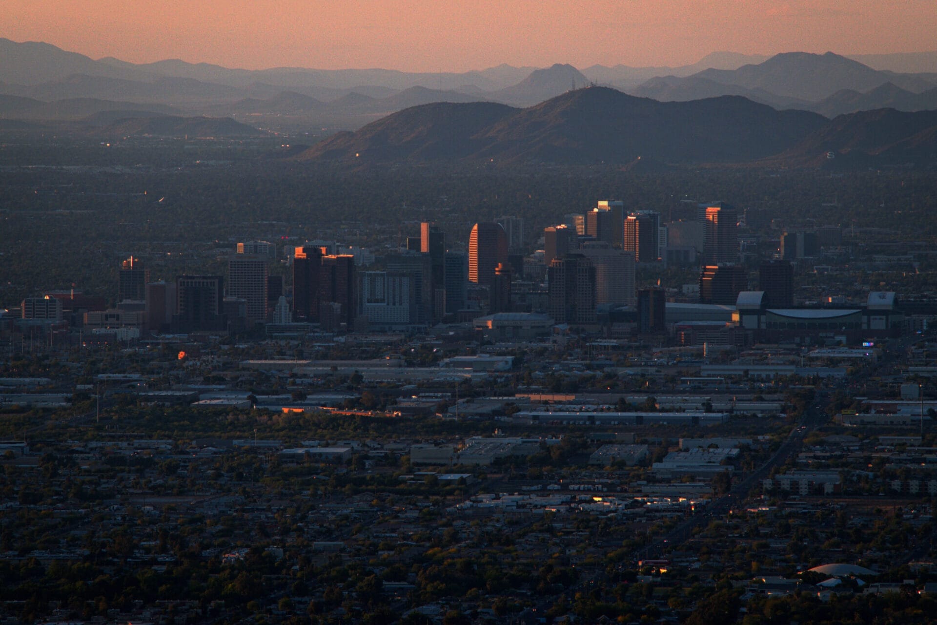 Aerial view of downtown Phoenix, Arizona, at sunset, showcasing the city's skyline, mountainous backdrop, and surrounding neighborhoods.