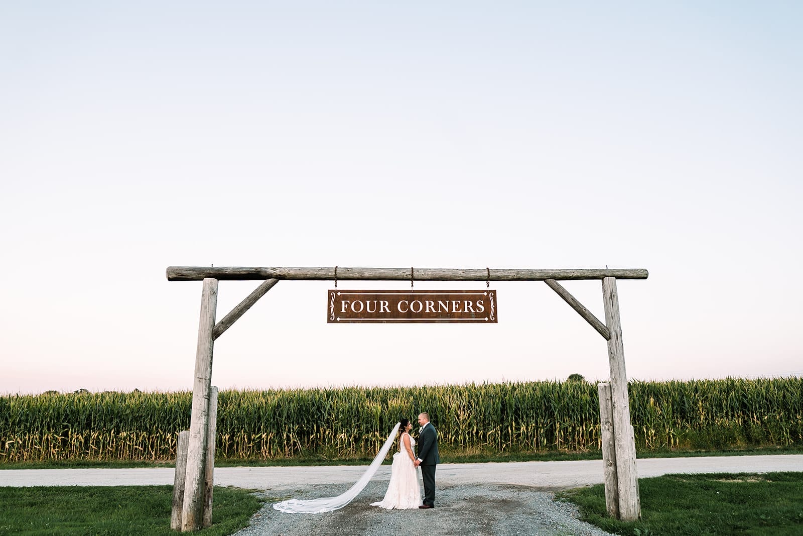 A newlywed couple stands under a wooden sign reading "FOUR CORNERS," surrounded by a cornfield at sunset. The bride wears a long veil and a white lace dress, while the groom is in a gray suit.