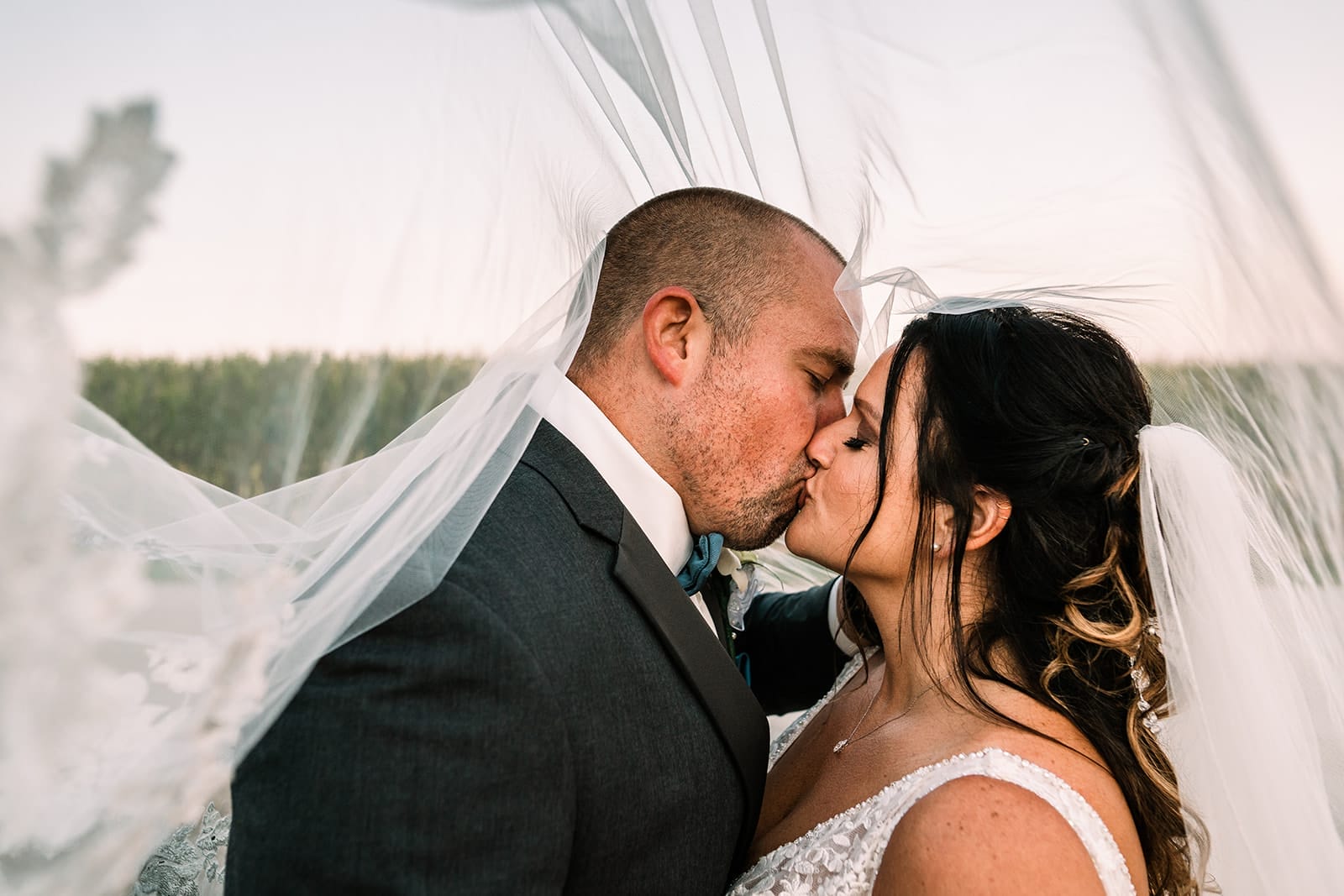 Bride and groom sharing a romantic kiss under a flowing veil at sunset, showcasing their elegant wedding attire and backdrop of nature.