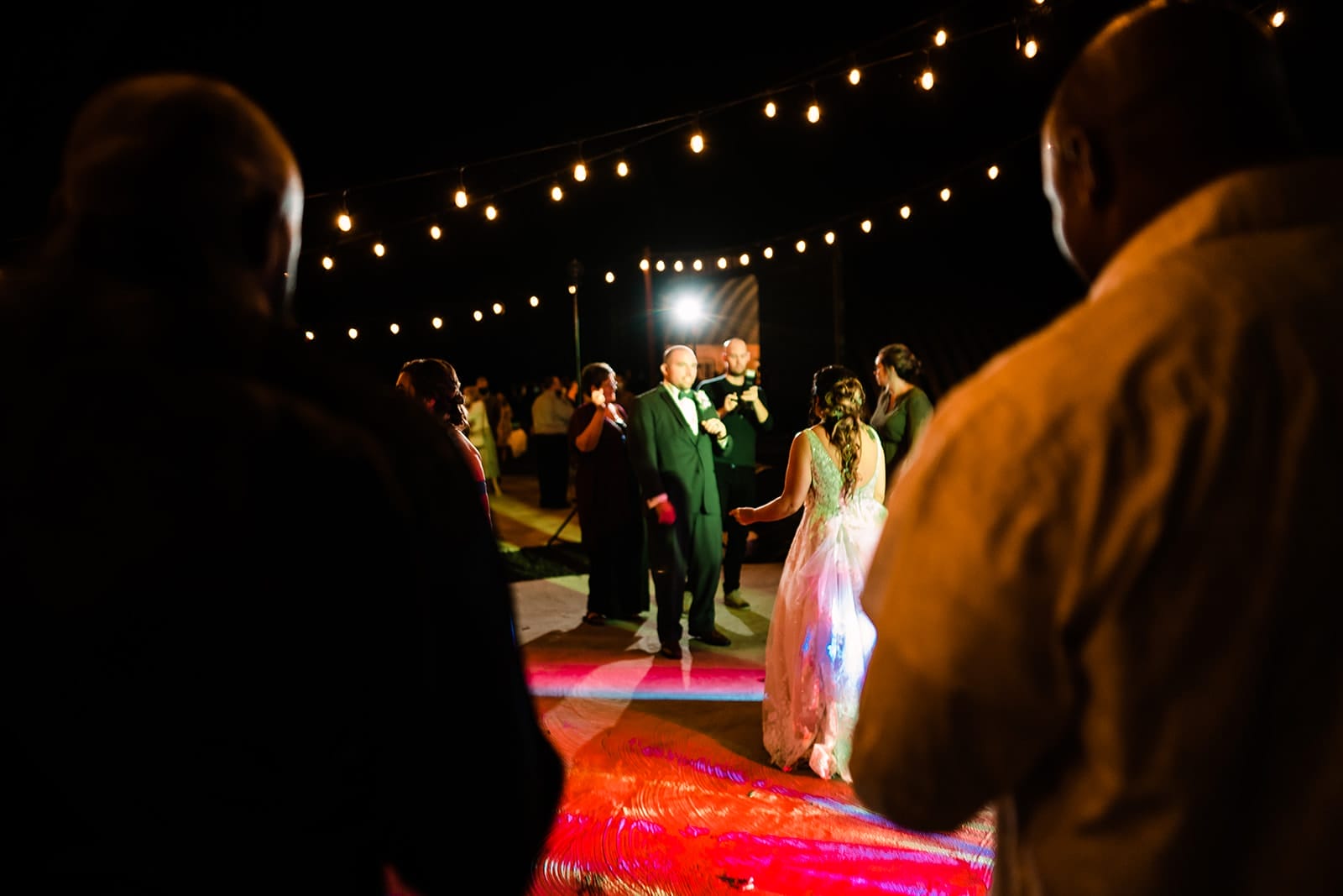 Guests dancing at a lively night event, illuminated by string lights, with a bride in a white gown enjoying the celebration in the foreground.