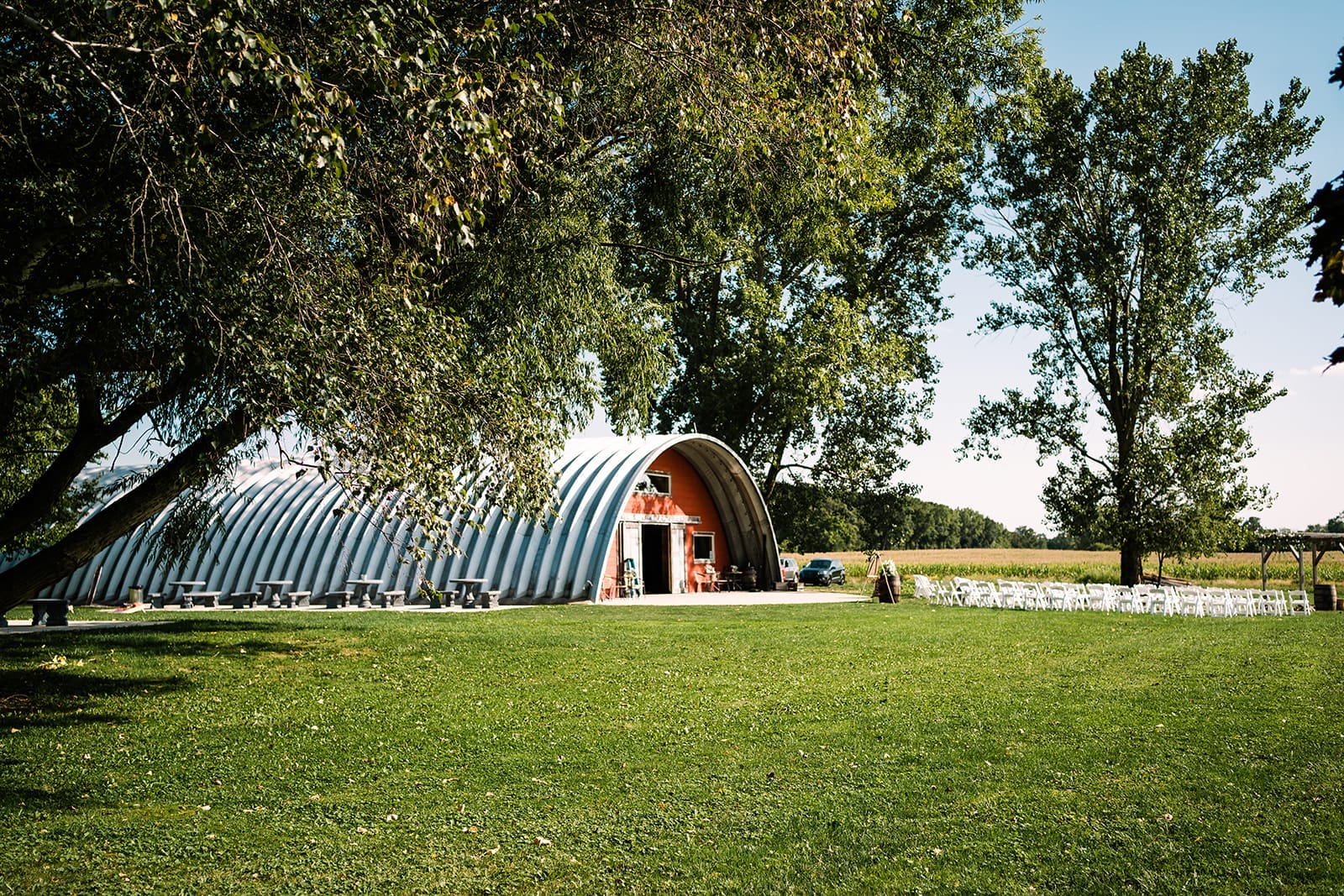 Modern barn venue surrounded by greenery, featuring a curved metal roof, outdoor seating, and a spacious lawn perfect for events.