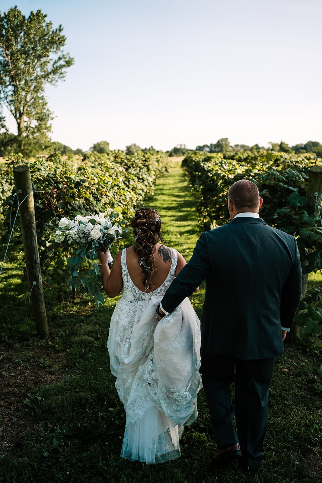 Bride and groom walking hand-in-hand through a vineyard, with lush green vines on either side. The bride wears an intricate lace wedding dress and holds a bouquet of white and green flowers, while the groom dons a formal suit. The scene captures a romantic outdoor wedding setting.
