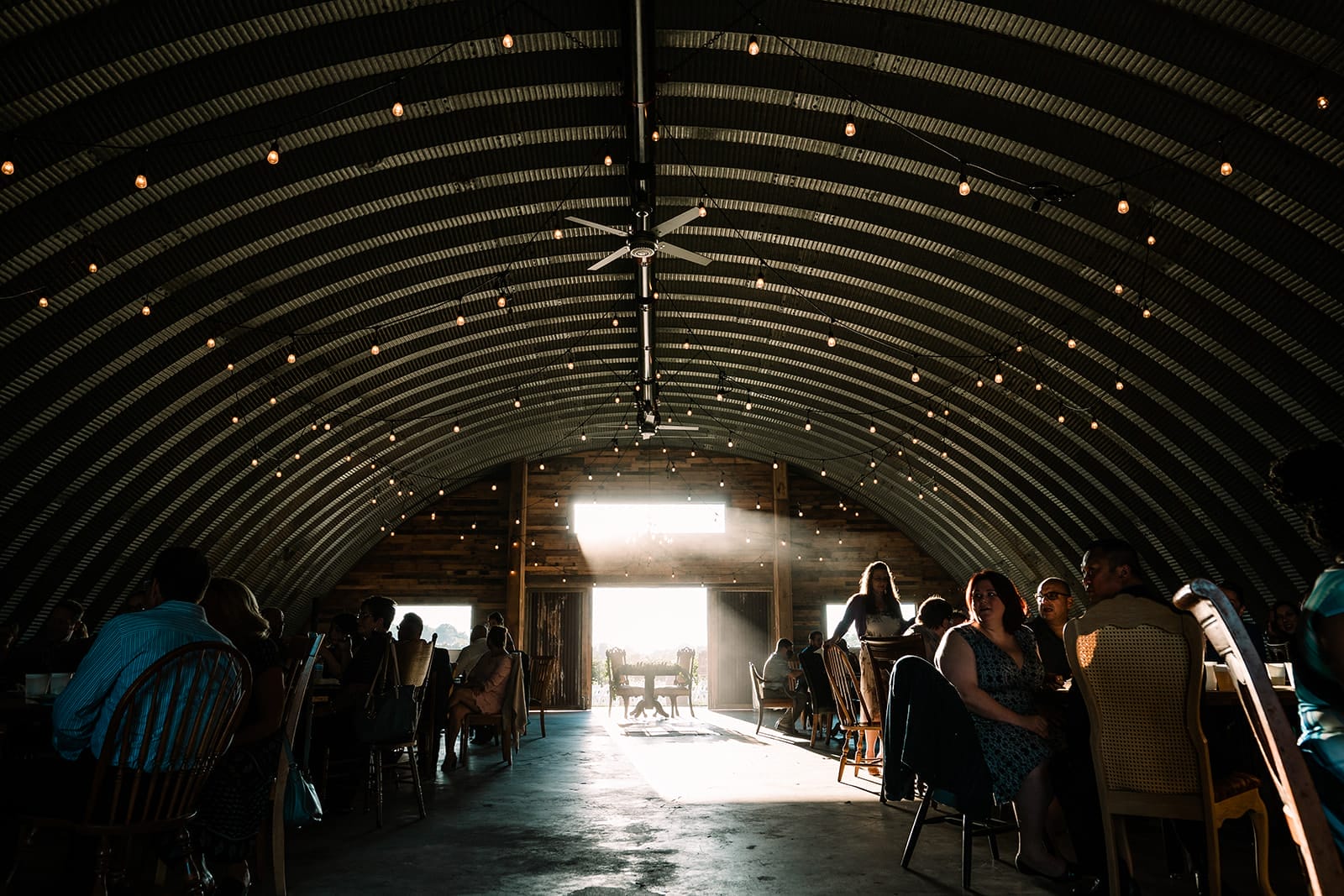 Interior view of a rustic event space featuring a curved metal roof and warm lighting, filled with seated guests enjoying a gathering, with sunlight streaming through an open doorway at the far end.