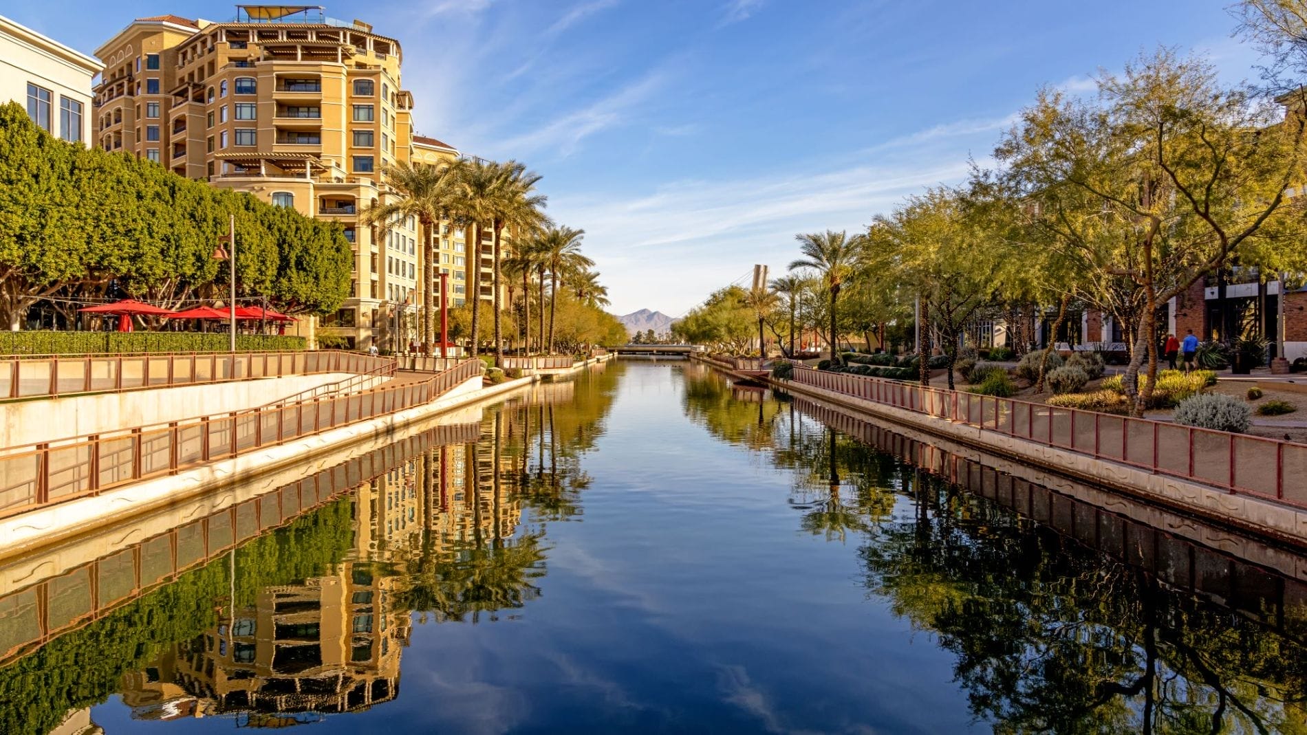 Scenic view of a calm canal lined with palm trees and modern buildings, reflecting the clear blue sky and nearby mountains in Scottsdale, Arizona.