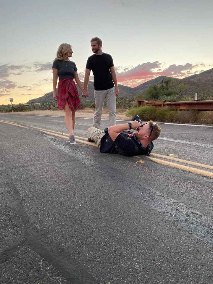 Couple holding hands on a road at sunset, with a photographer lying on the ground capturing their moment. Scenic mountains and a colorful sky in the background.